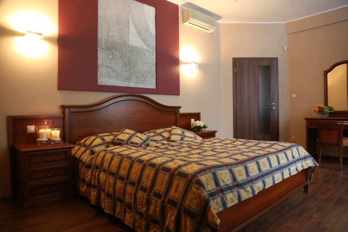 A bedroom with a wooden bed, a nightstand with a lamp, a dresser, a wall-mounted air conditioner, and a door.