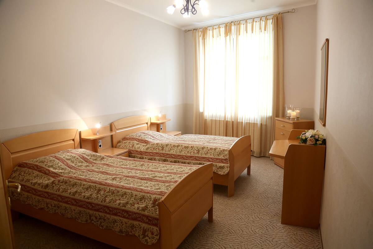 A hotel room with two wooden beds, a nightstand with a lamp, a dresser, and a window with curtains.