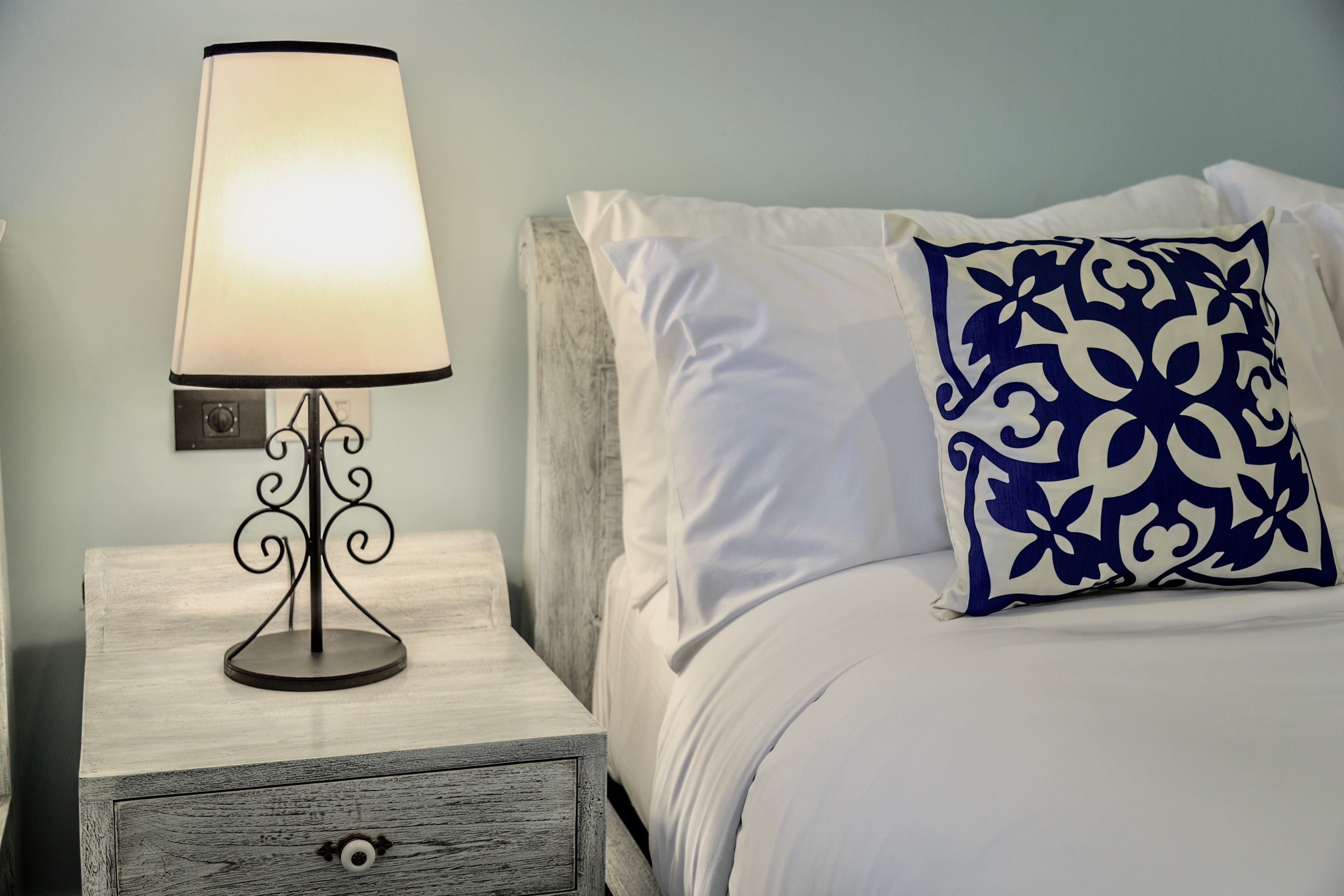 A neatly made bed with white bedding and a blue patterned pillow, a bedside lamp on a wooden nightstand, and a wall-mounted light switch.