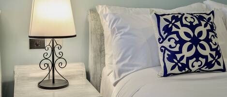 A neatly made bed with white bedding and a blue patterned pillow, a bedside lamp on a wooden nightstand, and a wall-mounted light switch.