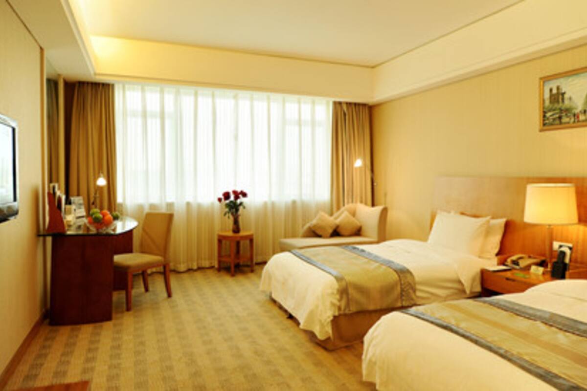 A hotel room with two beds, a desk with a chair, a small table with a vase of flowers, and a painting on the wall.