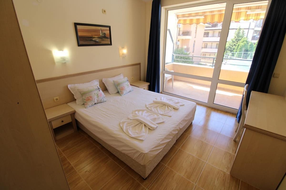 Ein Hotelzimmer mit Bett, Nachttisch, Kommode, einem Fenster mit Blick auf Gebäude und einem Wandbild.