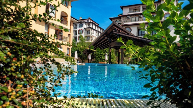A swimming pool with a wooden deck, surrounded by lush greenery and multi-story buildings with balconies. - Menada Paradise Dreams Apartments (Sveti Vlas)