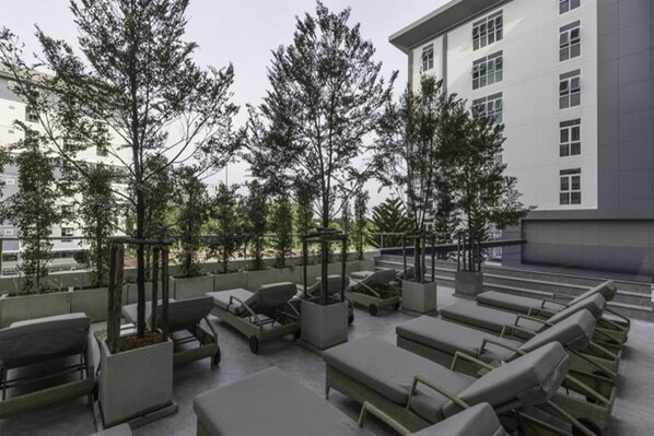 An outdoor seating area with lounge chairs and planters, surrounded by trees and a building in the background.