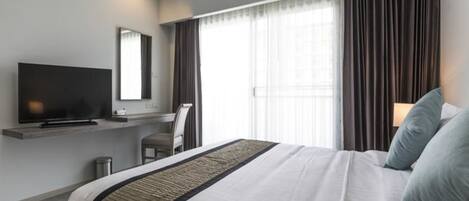 A hotel room with a bed, a TV on a stand, a desk, and a window with curtains.