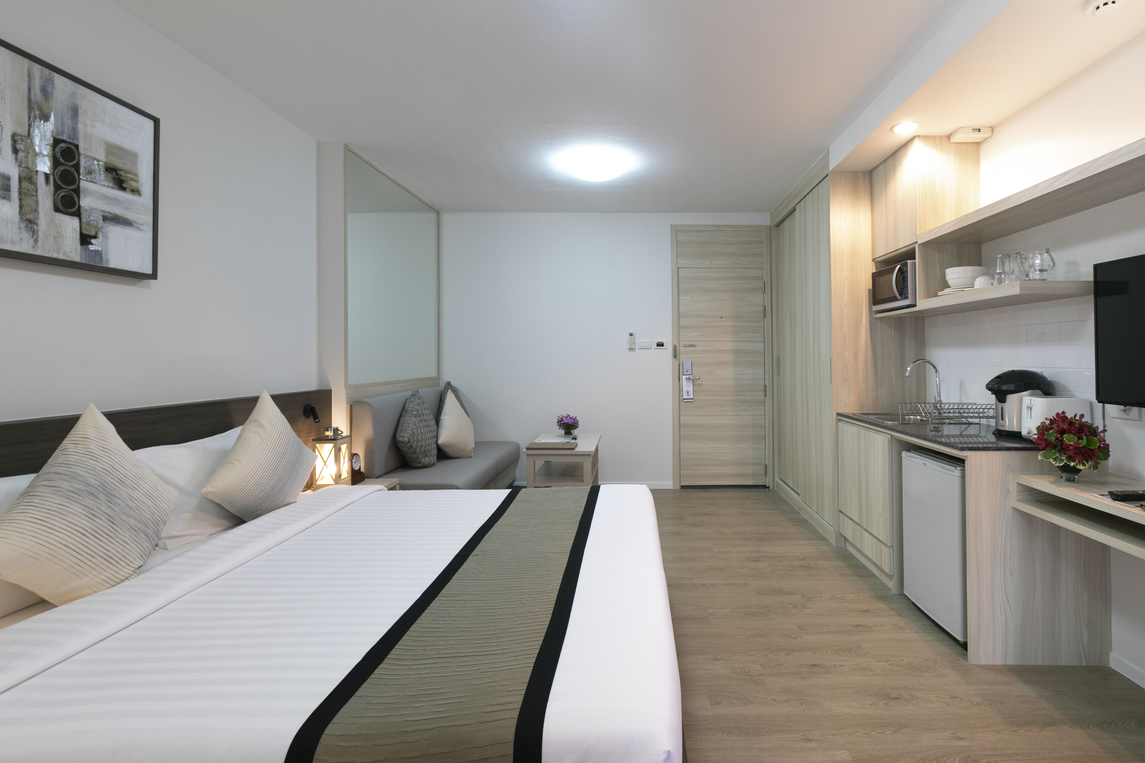 A modern hotel room with a bed, bedside table, lamp, and a view of the kitchen area.