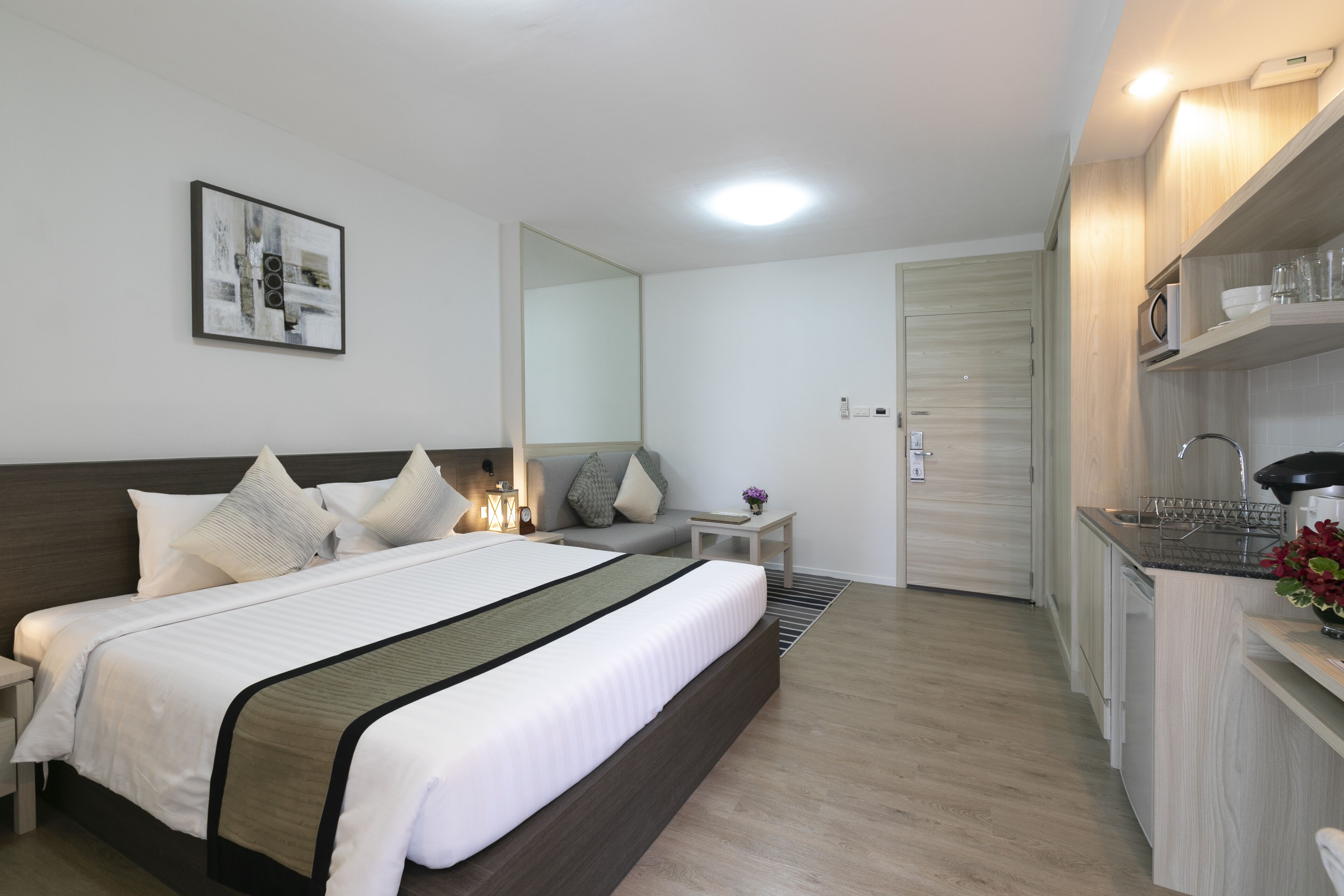 A modern hotel room with a large bed, a small seating area, a kitchenette, and a mirror.