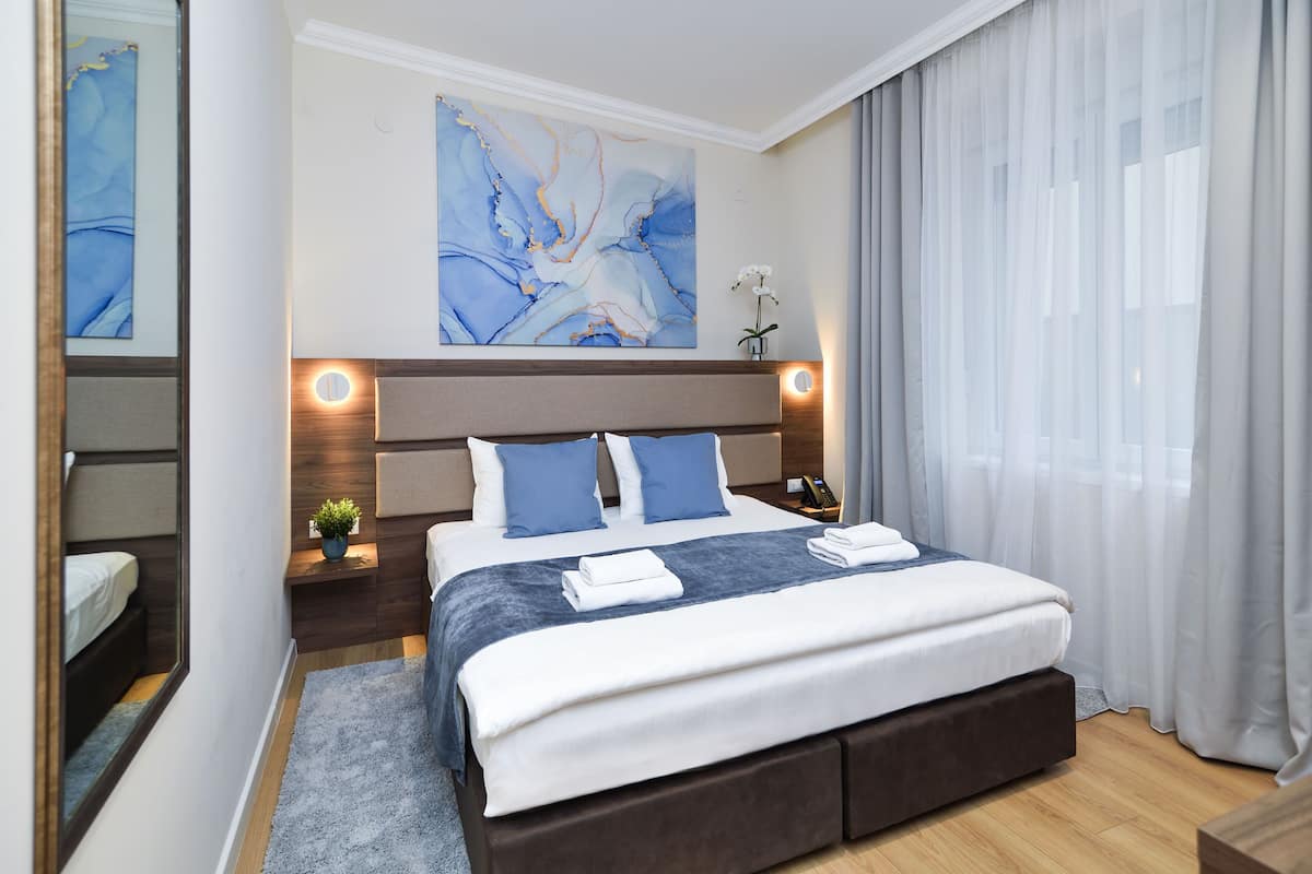 A modern hotel room with a large bed, a nightstand, a bedside table with a lamp, and a large abstract painting on the wall.