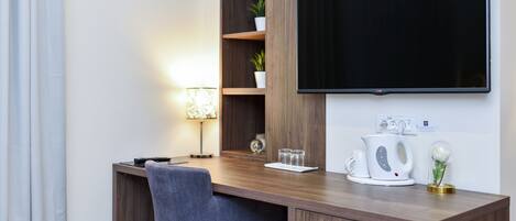 A modern workspace with a wooden desk, a chair, a flat-screen TV, a lamp, a kettle, and a small potted plant.