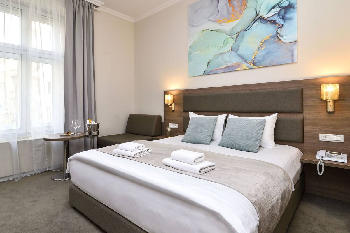 A hotel room with a large bed, two bedside tables with lamps, a small table with a bottle and glasses, and a painting on the wall.
