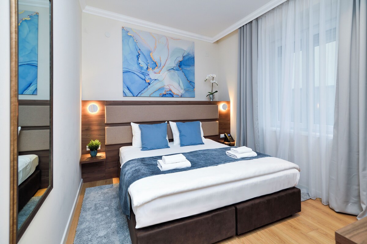 A modern hotel room with a large bed, a nightstand, a bedside table with a lamp, and a large abstract painting on the wall.