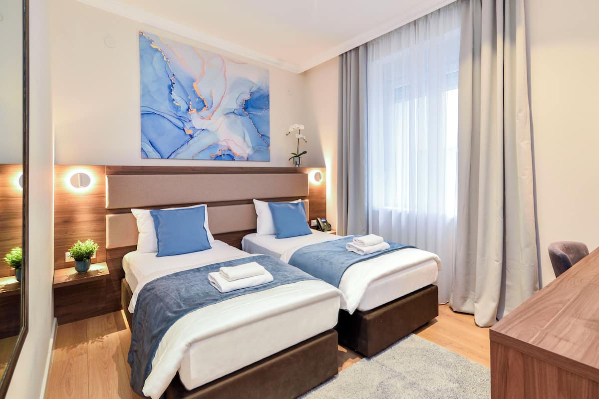 A hotel room with two beds, a large abstract painting, a bedside table with a lamp, and a view of the outdoors through sheer curtains.