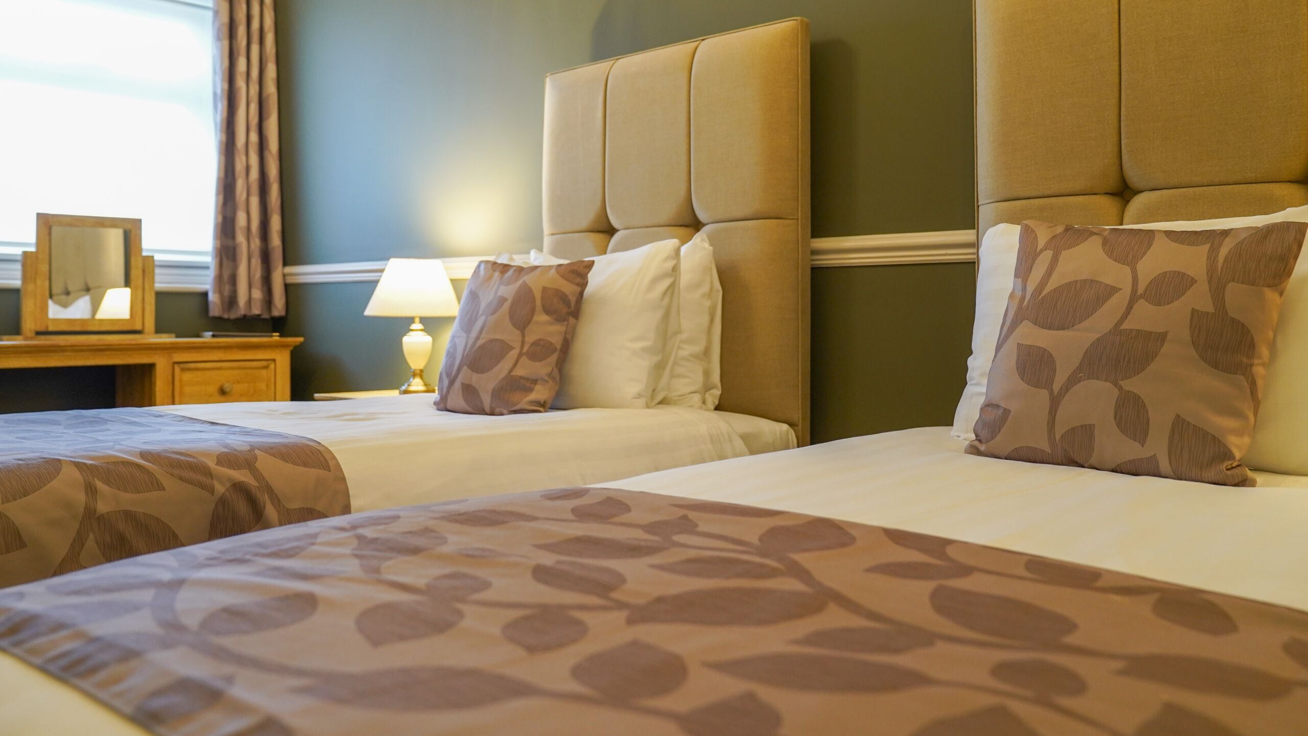 A hotel room with two beds, each with a headboard and a patterned bedspread. There is a nightstand with a lamp and a framed picture.