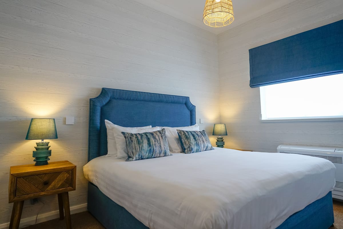 Ein Schlafzimmer mit einem großen Bett, zwei Nachttischlampen, einem Fenster mit blauer Jalousie und einem Holznachttisch.