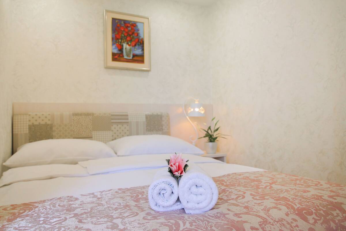 A neatly made bed with rolled towels and a flower arrangement, a bedside lamp, and a painting on the wall.