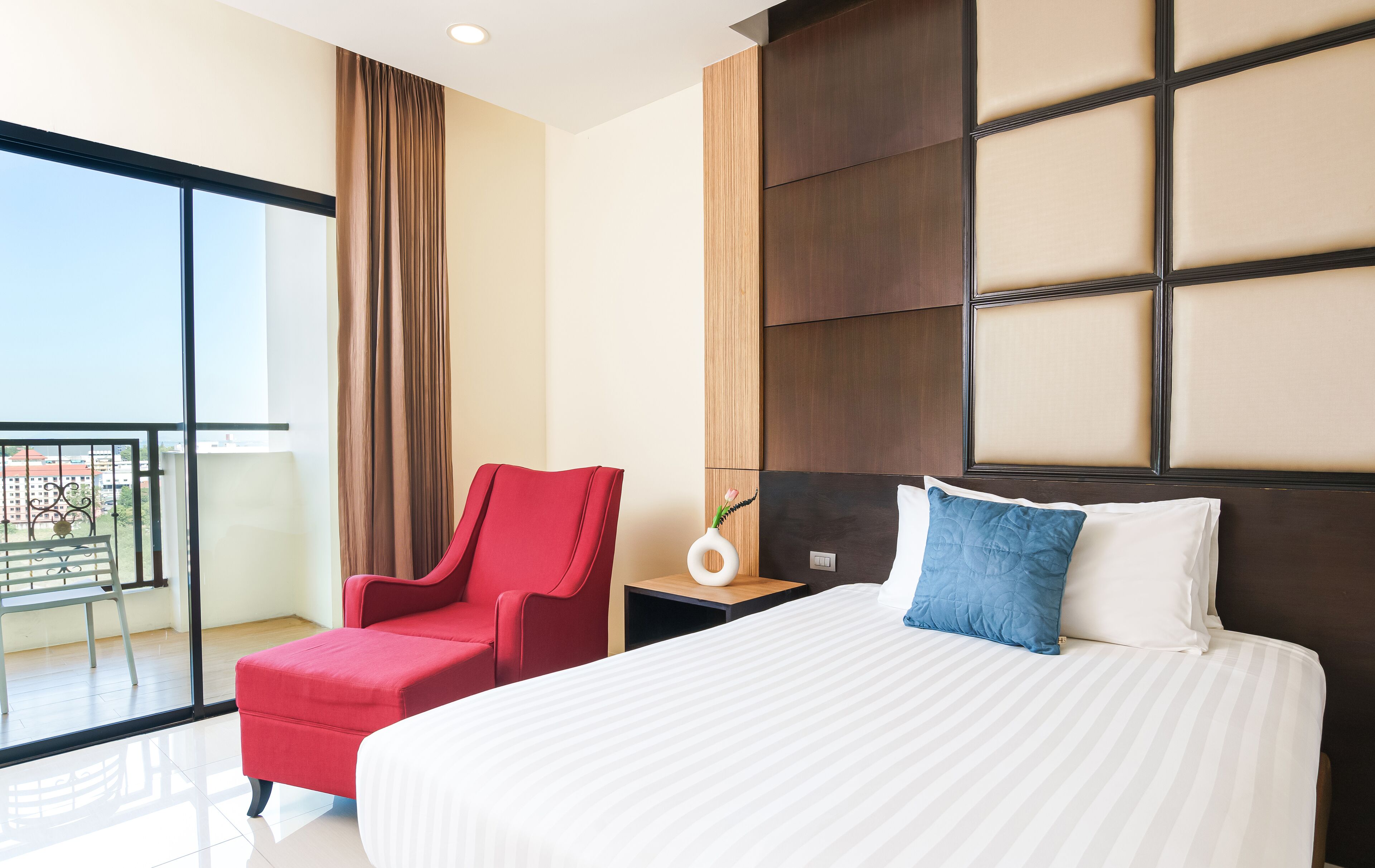 Quarto de hotel com uma cama grande, uma cadeira vermelha, uma mesa de cabeceira e vista para o exterior.