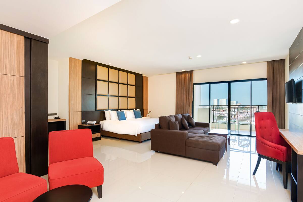 A modern hotel room with a large bed, a sofa, a red armchair, a small table, and a TV.