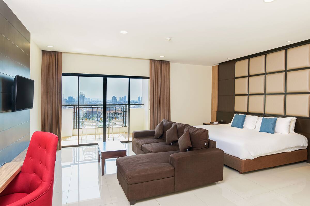 A modern hotel room with a large bed, a sofa, a red chair, and a TV on the wall.