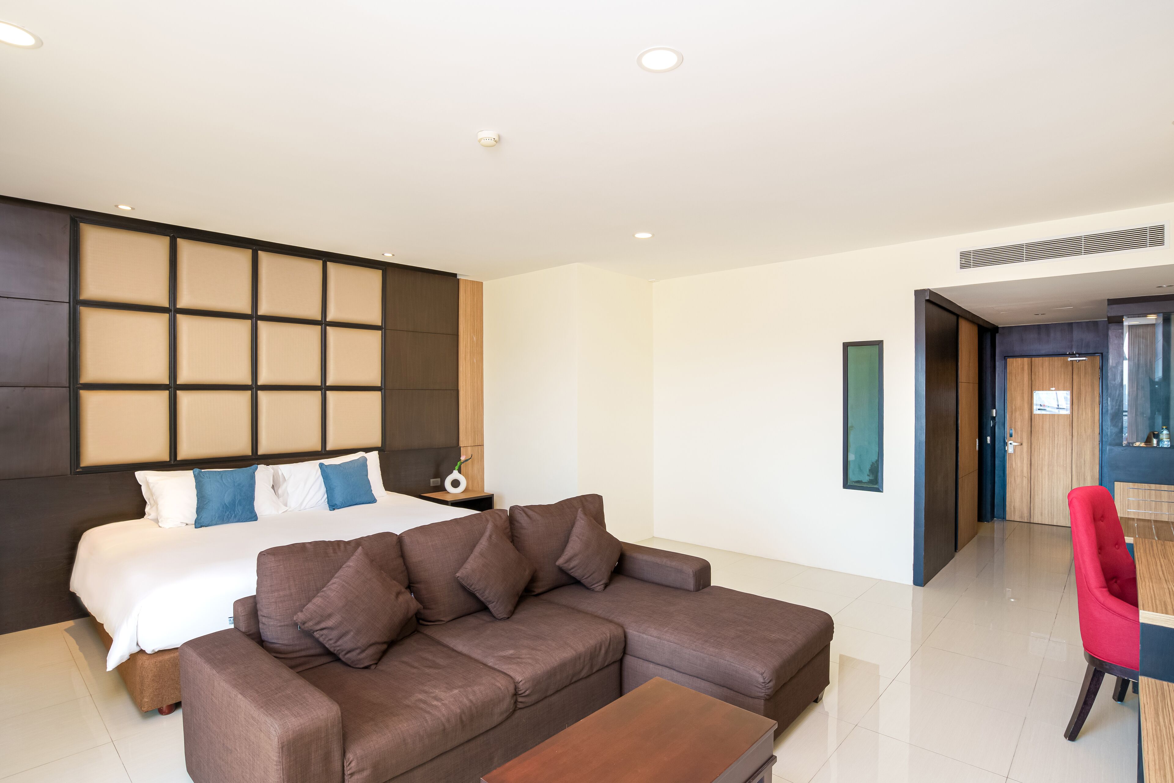 A modern hotel room with a large bed, a brown sofa, a wooden coffee table, and a red chair.