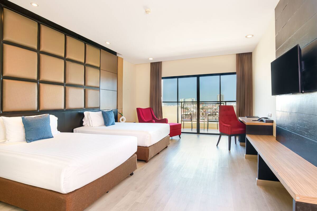 A hotel room with two beds, a desk, a chair, a TV, and a balcony with a city view.