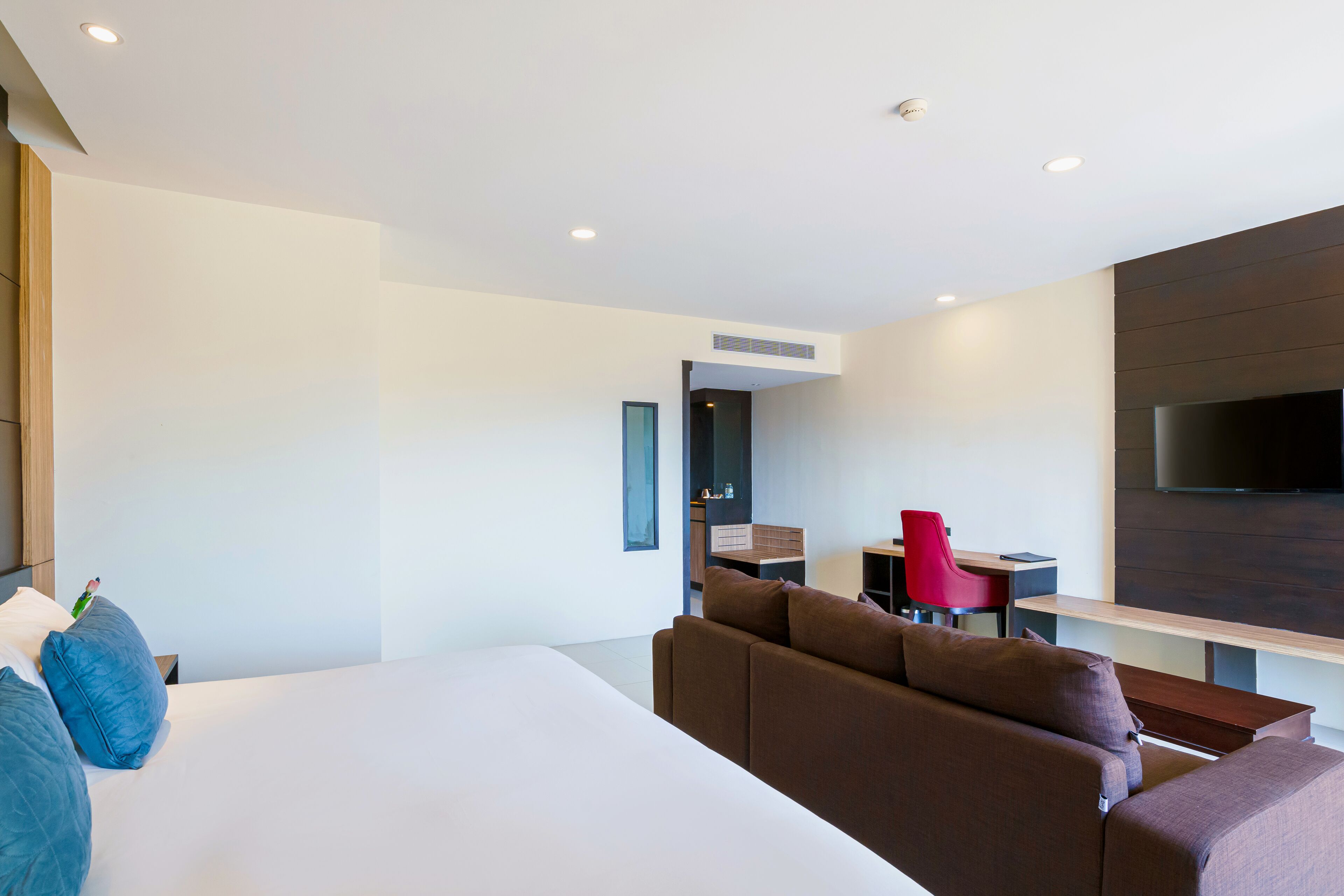 Une chambre d’hôtel moderne avec un grand lit, un canapé marron, un fauteuil rouge, un bureau et un téléviseur à écran plat fixé au mur.