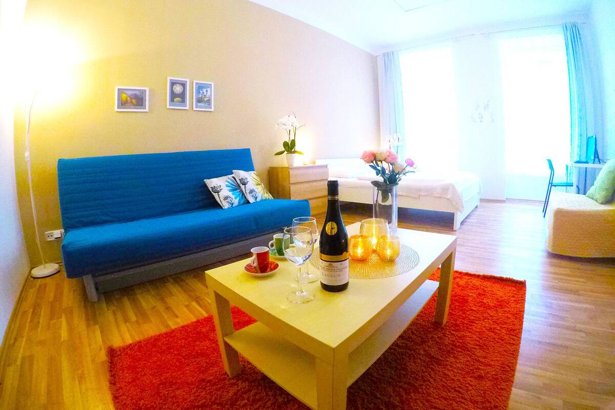 A modern living room with a blue sofa, a wooden coffee table with a bottle of wine, glasses, and candles, and a red rug.