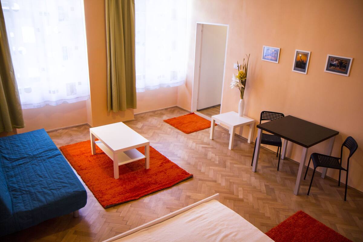 A room with a bed, a desk, a chair, and a small table. There is a window with curtains, a mirror, and a vase with flowers.