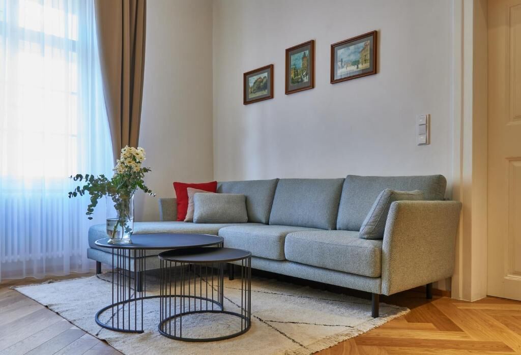 Een woonkamer met een grijze bank, een ronde salontafel en een vaas met bloemen. Er hangen drie ingelijste foto’s aan de muur.