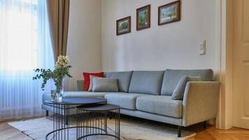 Een woonkamer met een grijze bank, een ronde salontafel en een vaas met bloemen. Er hangen drie ingelijste foto’s aan de muur.