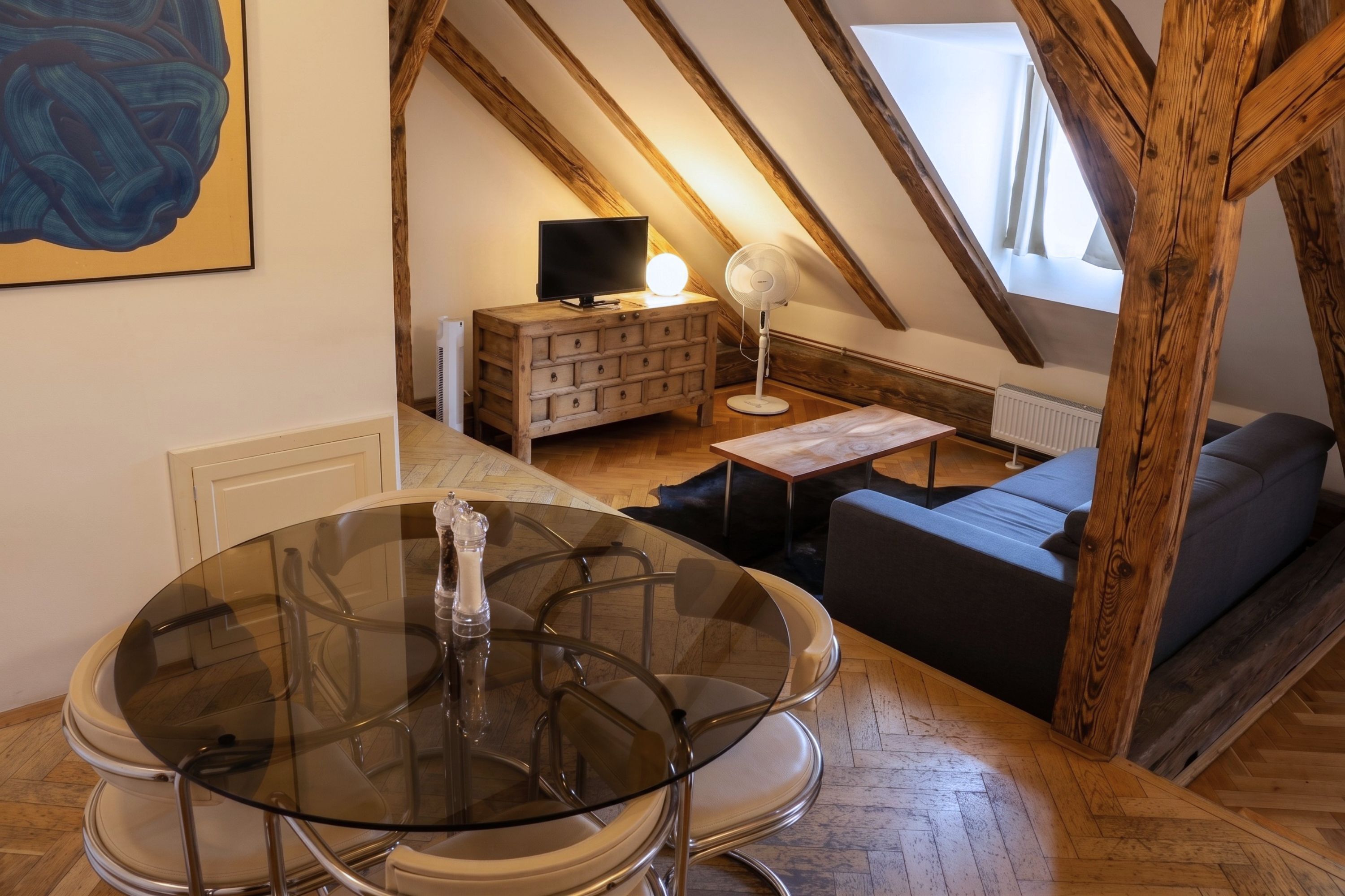 Un dormitorio tipo loft con una mesa de comedor de vidrio, un sofá, un televisor sobre un aparador de madera y un ventilador.