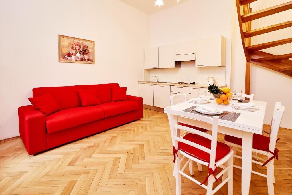 A compact living space with a red sofa, a dining table with white chairs, a kitchen area, and a wooden staircase.