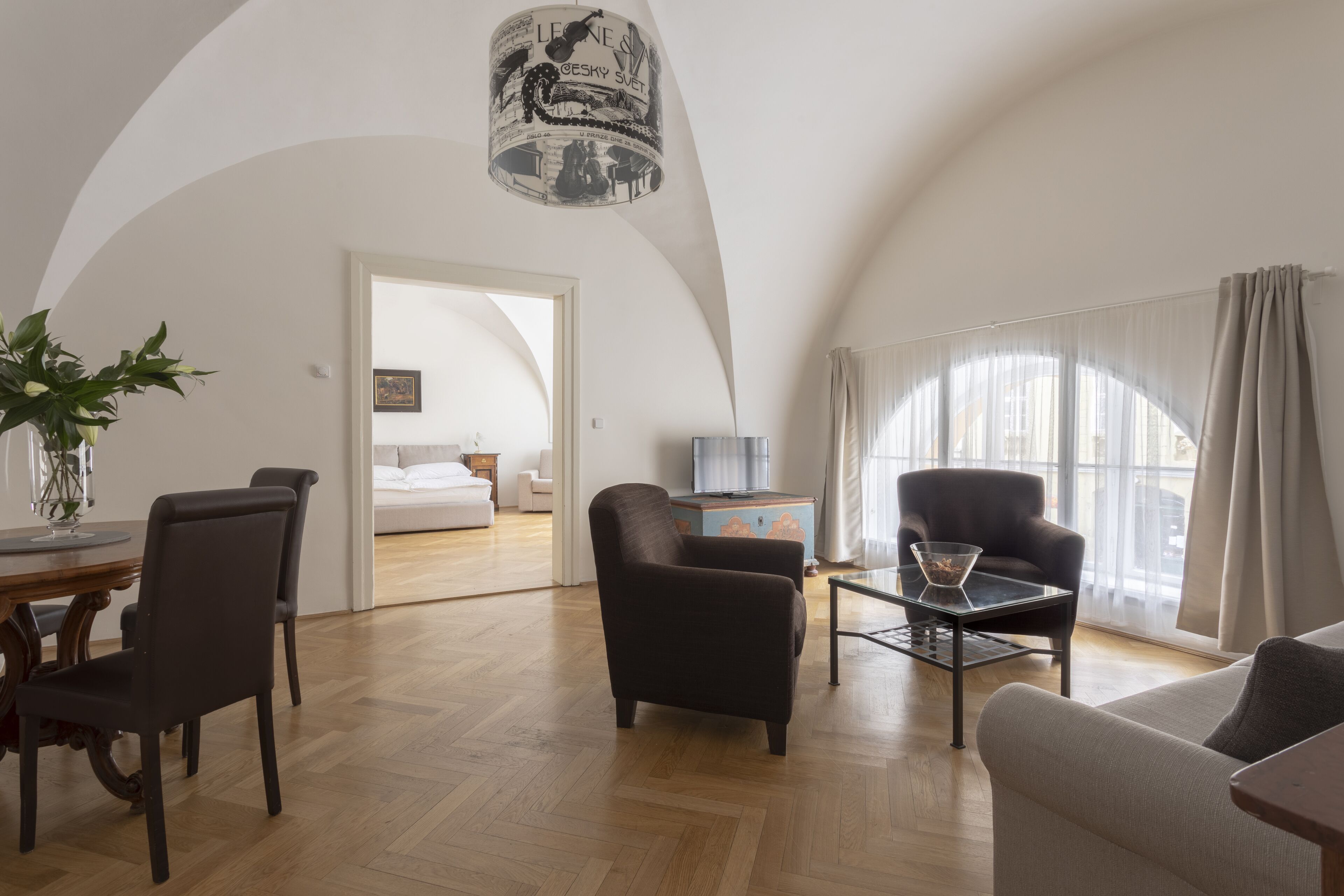 Amplia sala de estar con una mesa de comedor, sillas, un sofá y una mesa de centro. Cuenta con un amplio ventanal con cortinas, una lámpara de techo y una puerta que da a otra habitación.