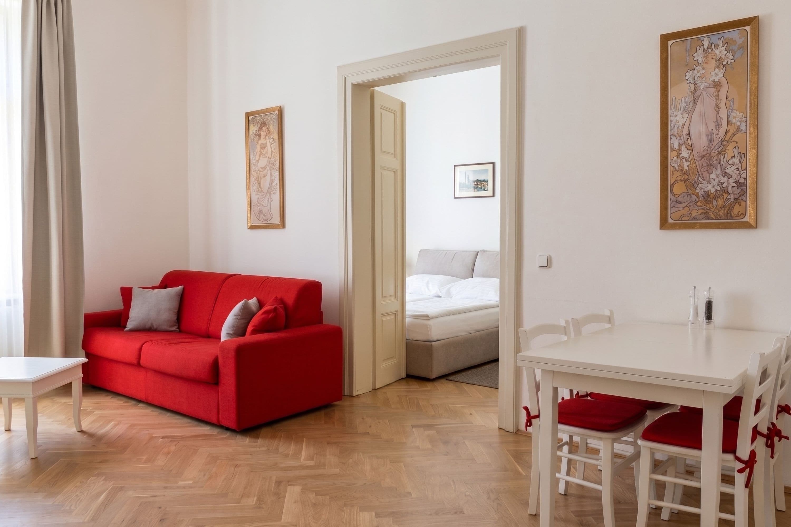Una habitación con un sofá rojo, una mesa de comedor blanca con cojines rojos, una cama blanca y una puerta que da a otra habitación.