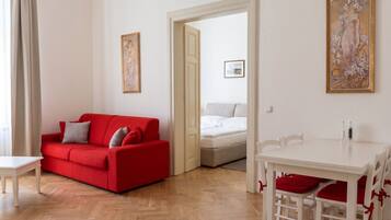 Una habitación con un sofá rojo, una mesa de comedor blanca con cojines rojos, una cama blanca y una puerta que da a otra habitación.