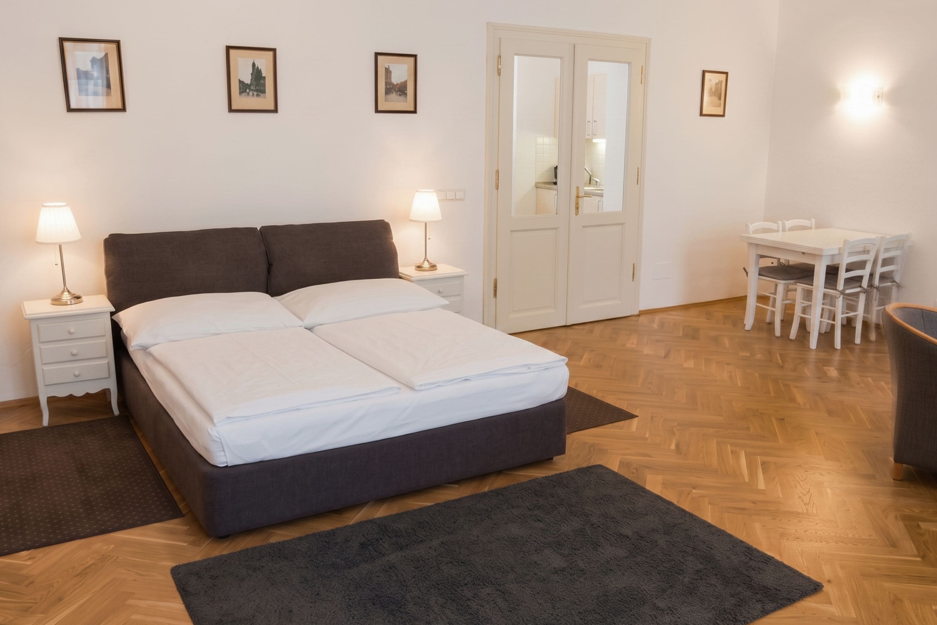 A bedroom with a bed, two bedside tables with lamps, a desk with chairs, and a door with a mirror.