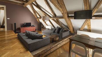 Un salón estilo loft con vigas de madera a la vista, un sofá seccional gris, una pequeña mesa de centro de madera y un comedor con una mesa sencilla y sillas.