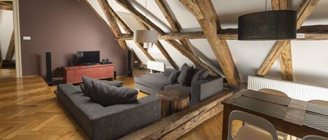Un salón estilo loft con vigas de madera a la vista, un sofá seccional gris, una pequeña mesa de centro de madera y un comedor con una mesa sencilla y sillas.