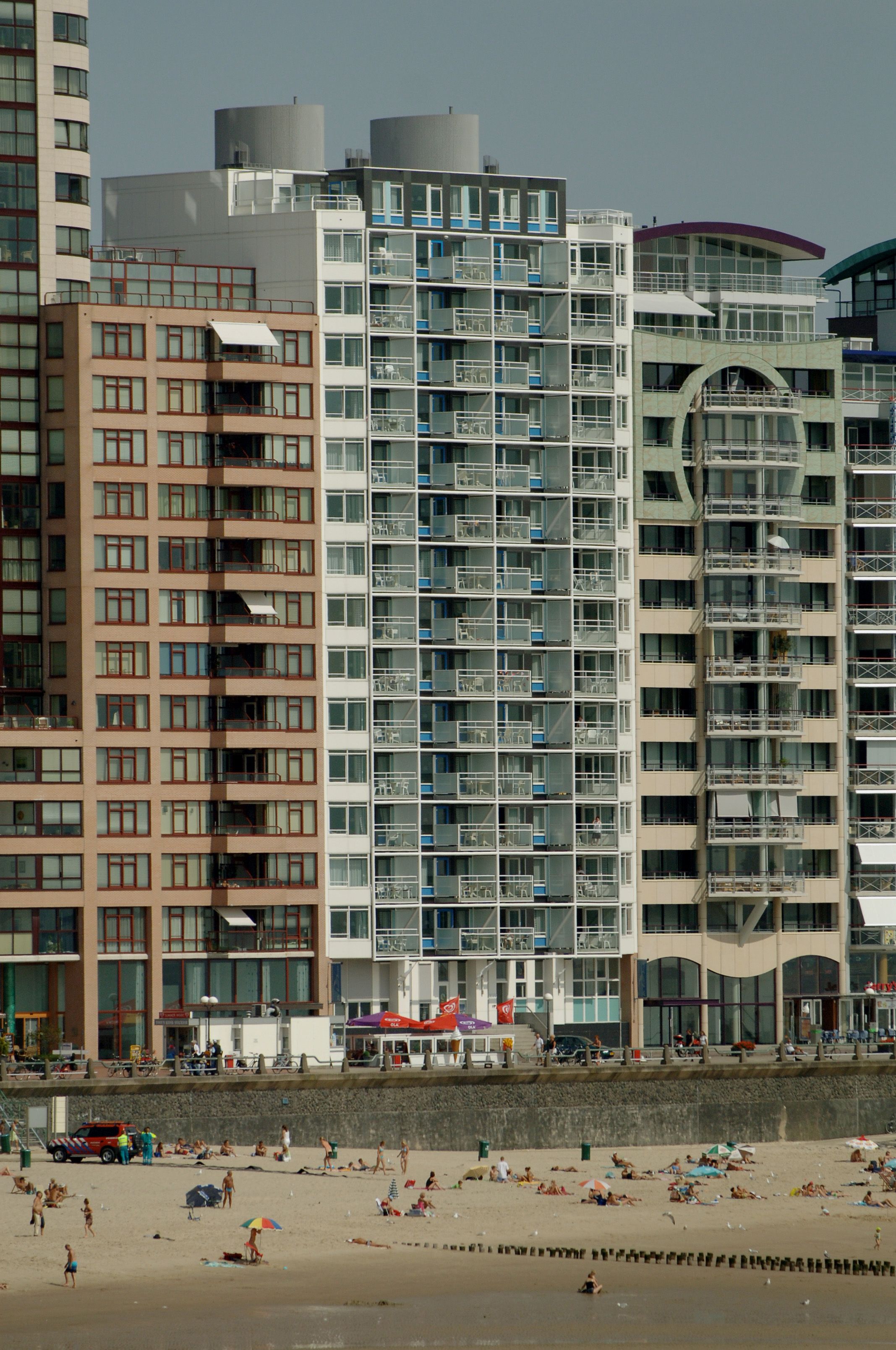 A high-rise hotel with multiple balconies, a beach in the foreground, and a clear sky.