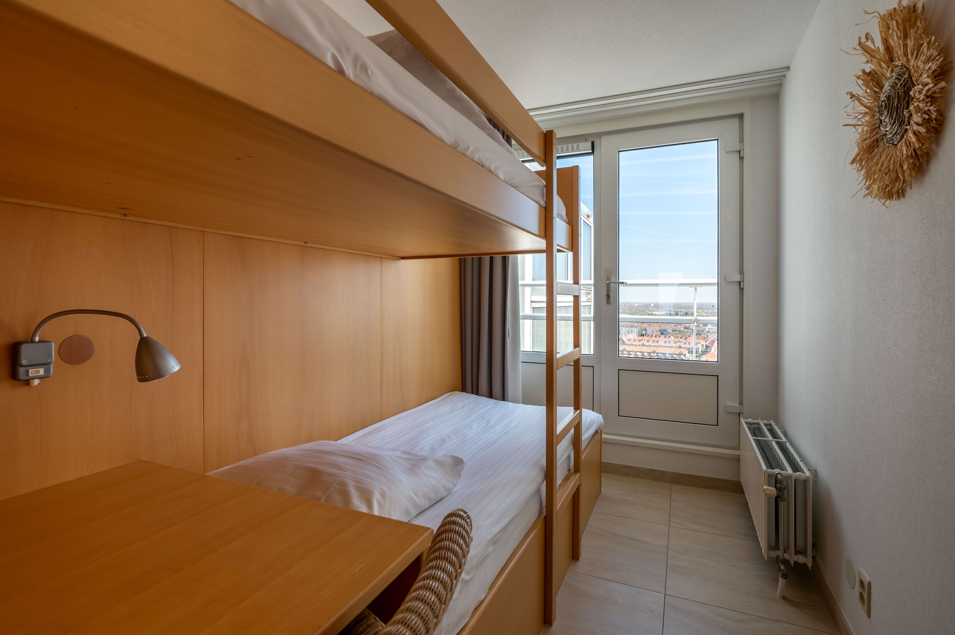 A compact room with a bunk bed, a desk, a lamp, and a view of the city through a window.