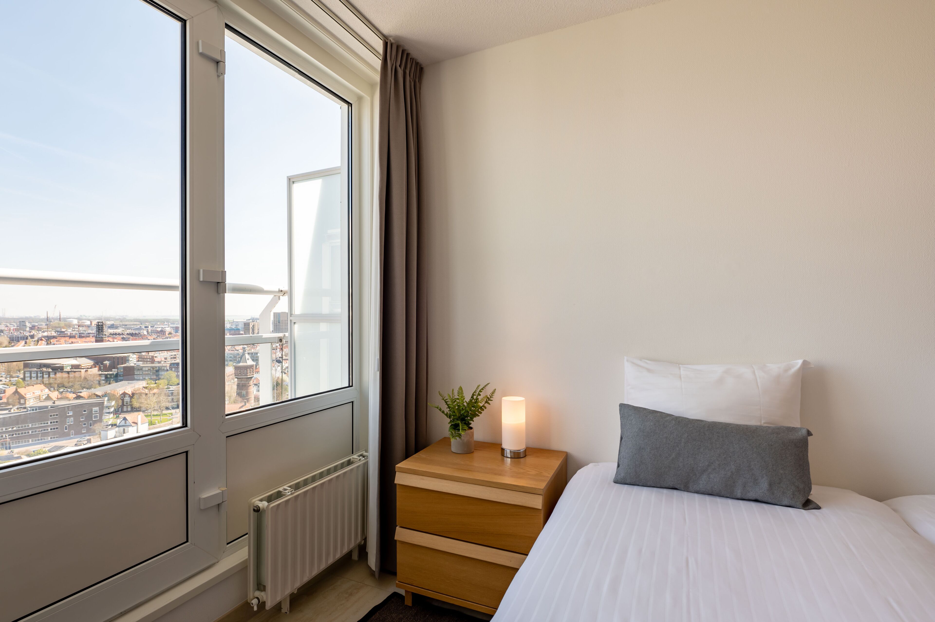 A hotel room with a large window, a bedside table with a lit candle, a bed with white bedding, and a view of a cityscape.