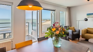 A room with a dining table, a vase of flowers, a sofa, and a view of the beach through large windows.