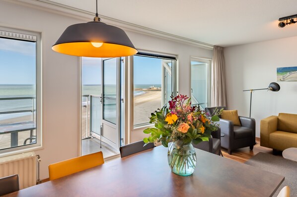 A room with a dining table, a vase of flowers, a sofa, and a view of the beach through large windows.