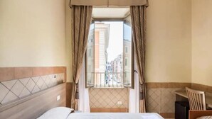 A hotel room with a bed, a desk, and a view of the city through a window. - Alex Romano (Rome)