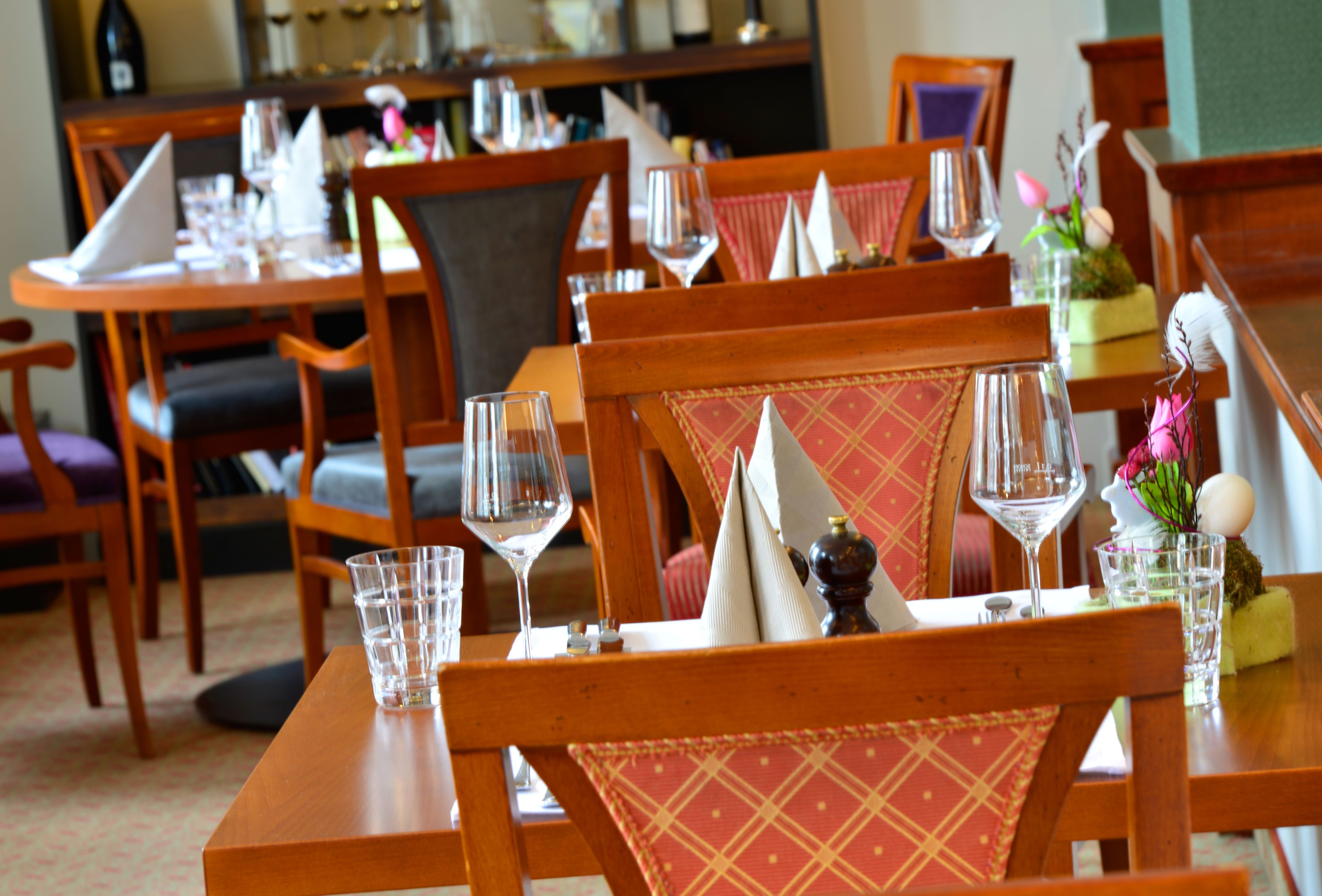 A dining area with wooden tables and chairs, each set with a wine glass, napkin, and a small decorative item.