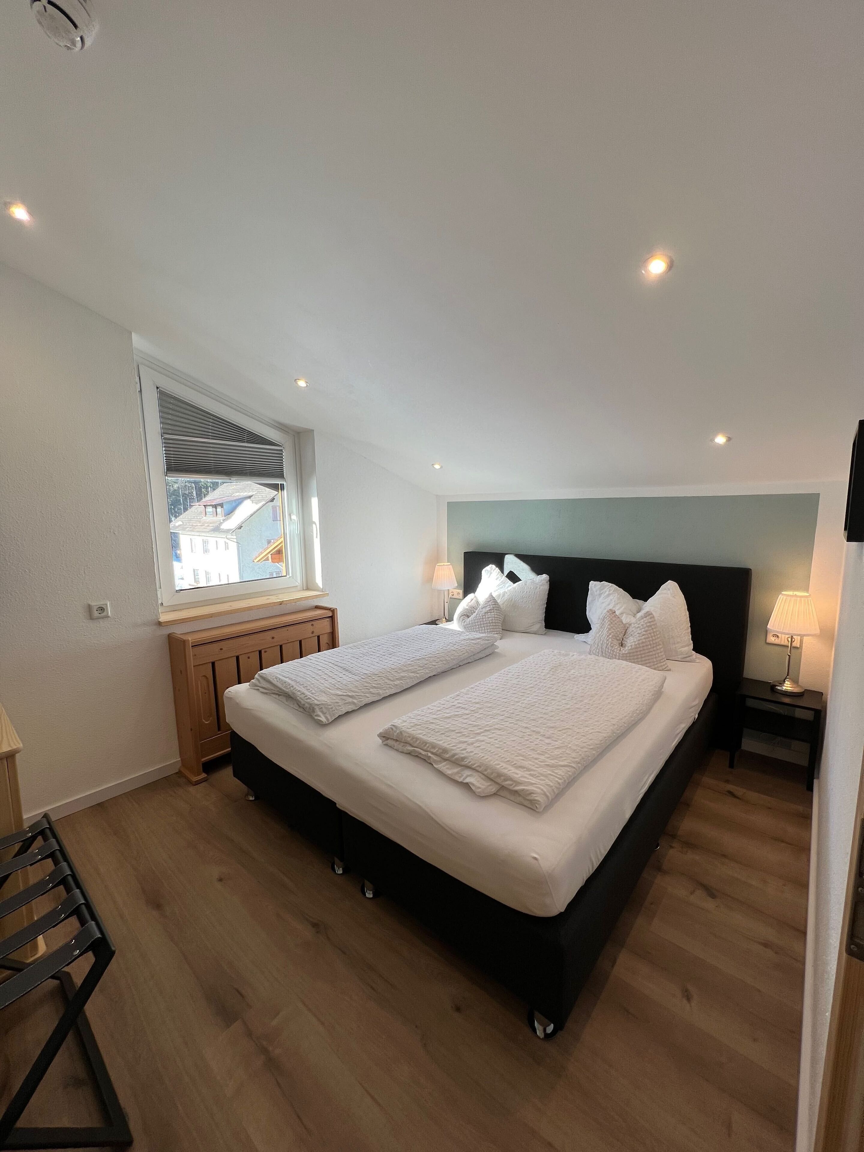A bedroom with a large bed, a window with blinds, a wooden nightstand, and a lamp.