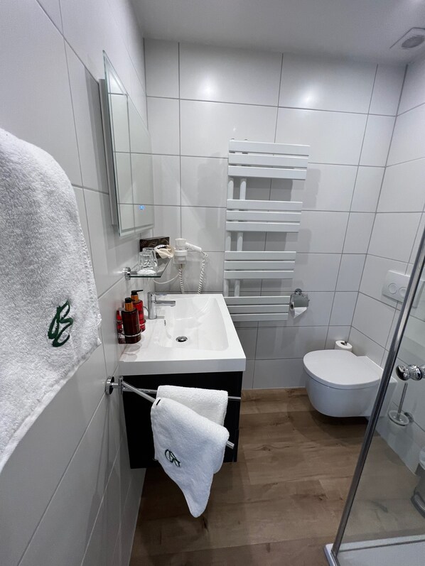 A modern bathroom with a white sink, mirror, and toilet. There are towels hanging on the wall and a glass shower door. - Hotel Berghof (Berg im Drautal)