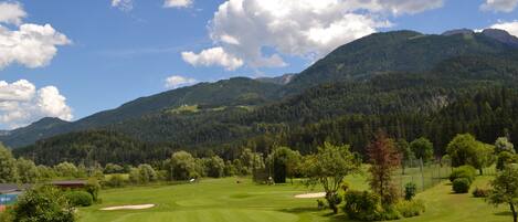 A golf course with a clear sky, green grass, and surrounding forested hills.