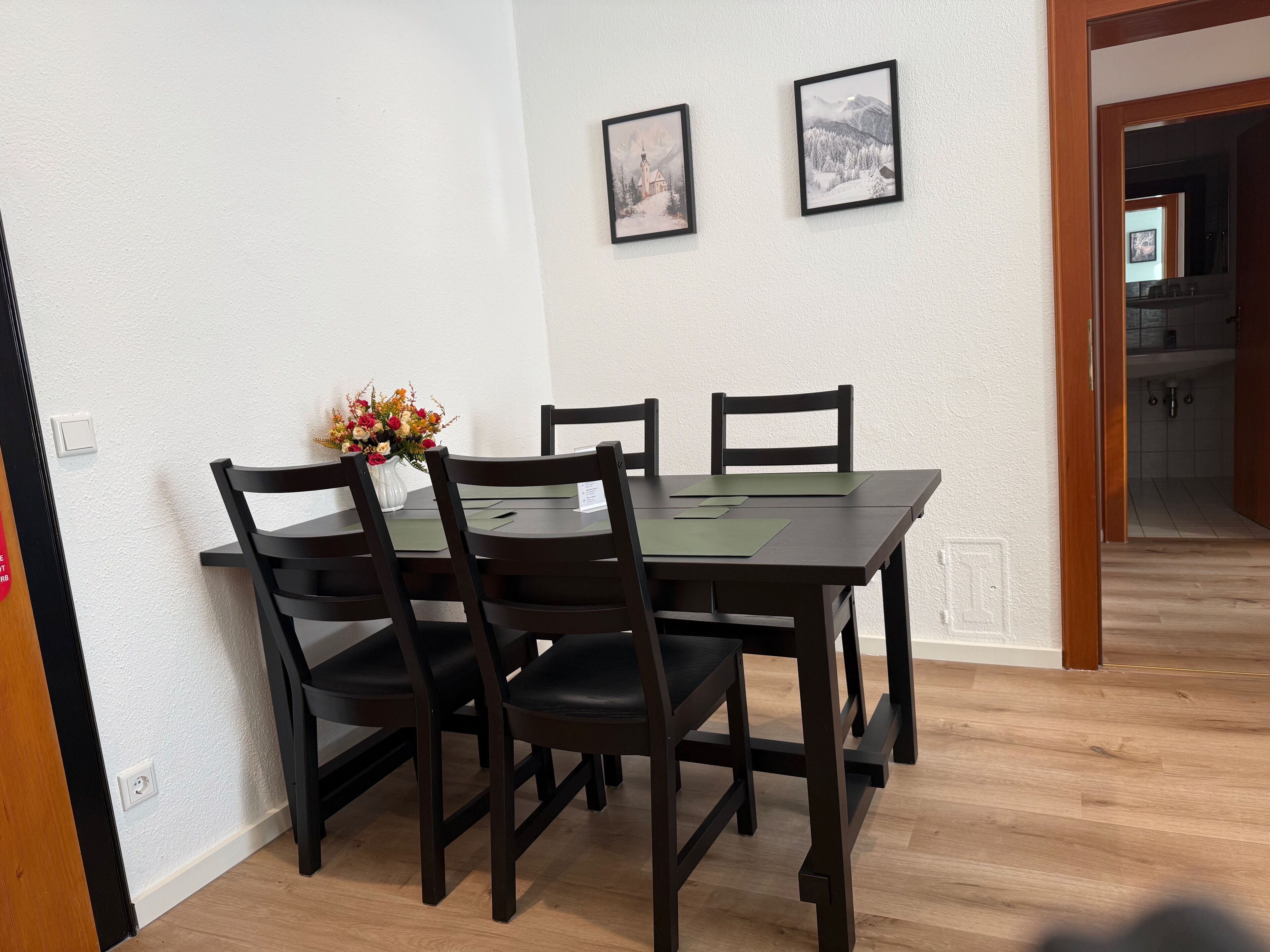 A dining area with a wooden table and chairs, a vase with flowers, and two framed pictures on the wall.
