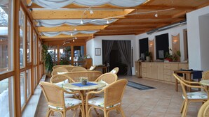 A dining area with wicker furniture, a tiled floor, and a kitchen area with wooden cabinets and a countertop. - Hotel Berghof (Berg im Drautal)