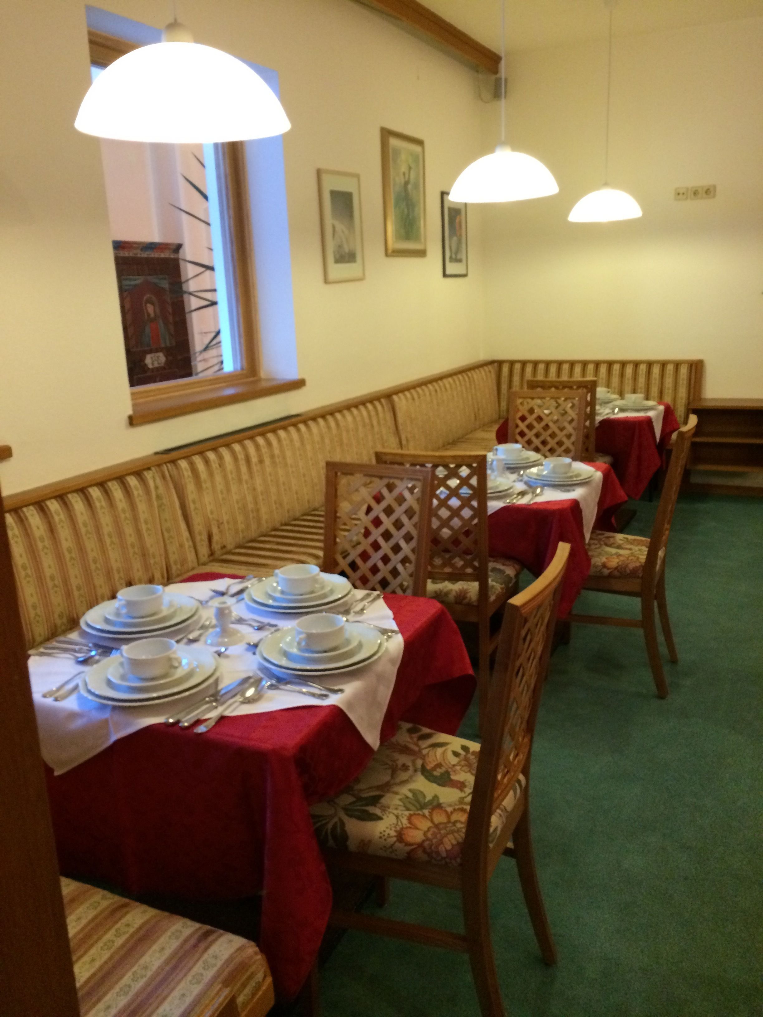 A dining area with tables set for four, each with a red tablecloth, white plates, and silverware. There are wooden chairs with floral upholstery. The room has a green carpet, wooden paneling on the walls, and a window with a view of a building.